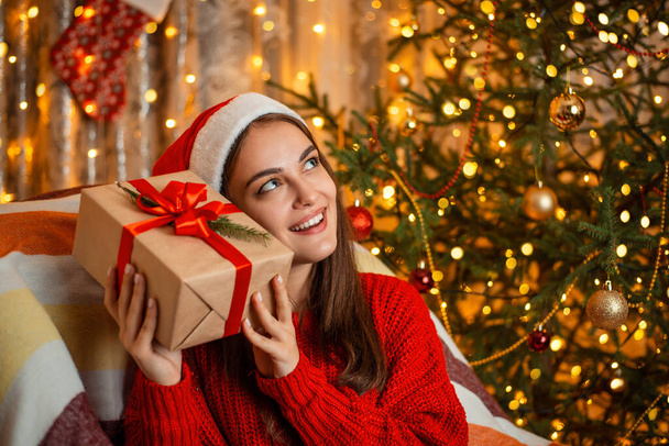 Νεαρό όμορφο κορίτσι σε καλή διάθεση προσπαθεί να μαντέψει τι περιέχει το κουτί δώρου. Χριστουγεννιάτικο δέντρο και ζεστό κίτρινο φως γιρλάντες στο παρασκήνιο, εορταστική ατμόσφαιρα. - Φωτογραφία, εικόνα