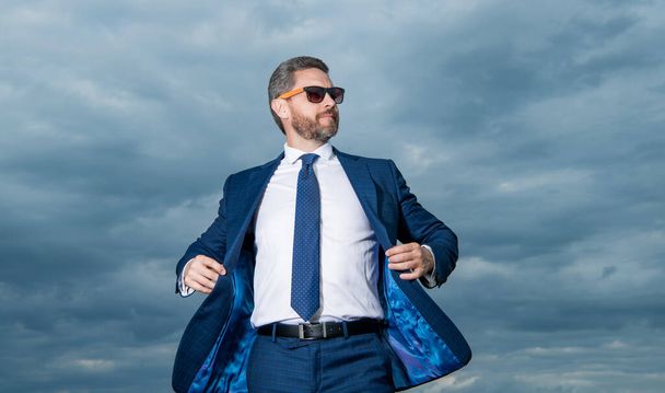 Guy wearing open suit jacket fluttering in wind. Grizzled guy in sunglasses. Professional guy sky background. - Photo, Image