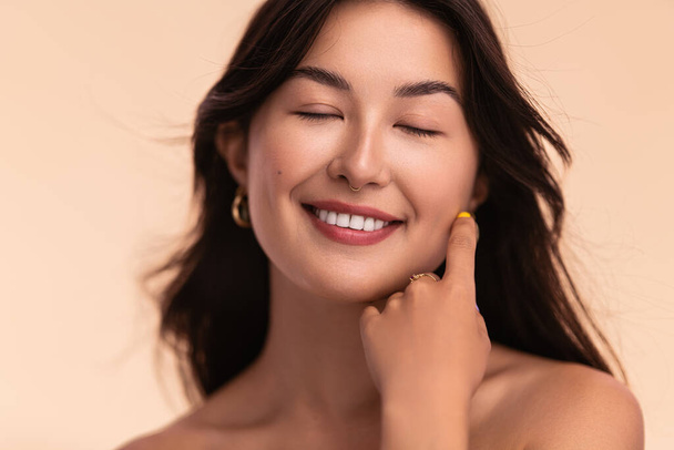 Veselý asijský ženský model s hladkou pletí a hnědé vlasy dotýkající se obličeje pro koncepci krásy rutiny na béžovém pozadí - Fotografie, Obrázek