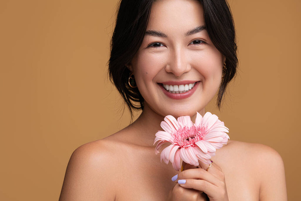 Cheerful young Asian female with bare shoulders holding pink gerbera flower and looking at camera with smile against brown background - Foto, imagen