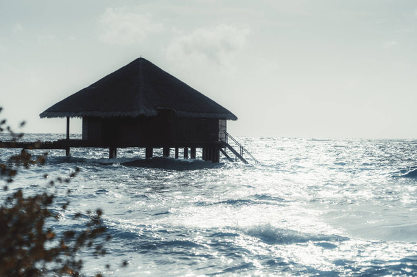 A bluish seascape with a silhouette of a bungalow with a triangle roof as a part of a group of cottages of a luxury Maldives island hotel on a warm sunny day surrounded by ocean waves - Φωτογραφία, εικόνα