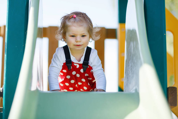 Мила дівчинка, яка грає на слайді на відкритому майданчику. Красива дитина в червоних гумових штанях розважається в сонячний теплий літній день. Дитина ковзає вниз
. - Фото, зображення
