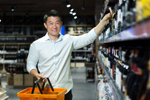 A young handsome Asian man businessman in a white shirt chooses wine in the alcohol section of a supermarket, takes one bottle. He holds a product basket in his hand, looks at the camera, and smiles. - Fotó, kép