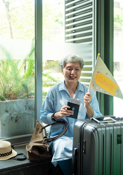 Muotokuva vanhempi aikuinen vanhusten aasialainen nainen 60s hymyillen istuu matkalaukku matkalaukku ja tilalla opastettu lippu napa matkustaa käsite. - Valokuva, kuva