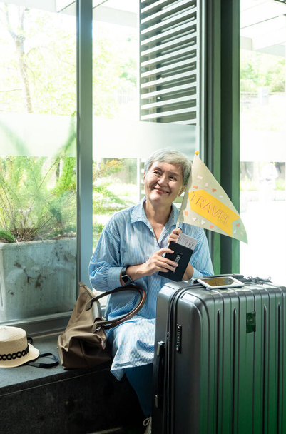 Portrait de femme asiatique âgée âgée des années 60 souriant assis avec sac à bagages valise et tenant un mât de drapeau guidé pour le concept de voyage. - Photo, image