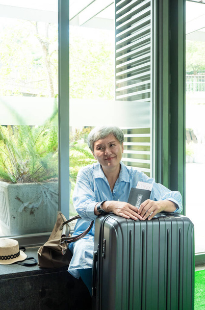 Portret starszej osoby w podeszłym wieku Azjatki 60-letnia uśmiechnięta kobieta siedząca z walizką na bagaż i posiadająca paszport z biletem na podróż. - Zdjęcie, obraz