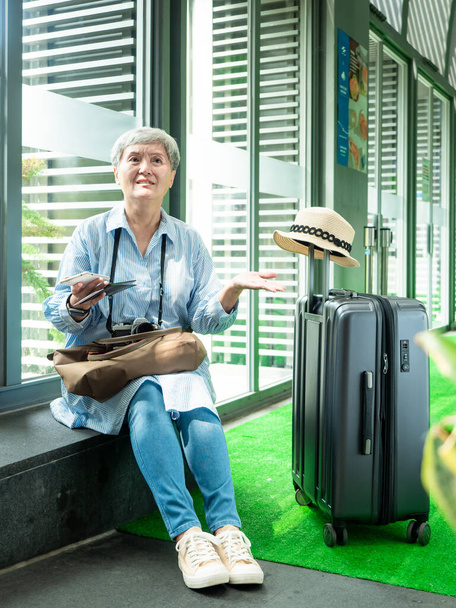 Портрет пожилой женщины 60 лет, улыбающейся сидя с чемоданом и паспортом с билетом на концепцию путешествия. - Фото, изображение