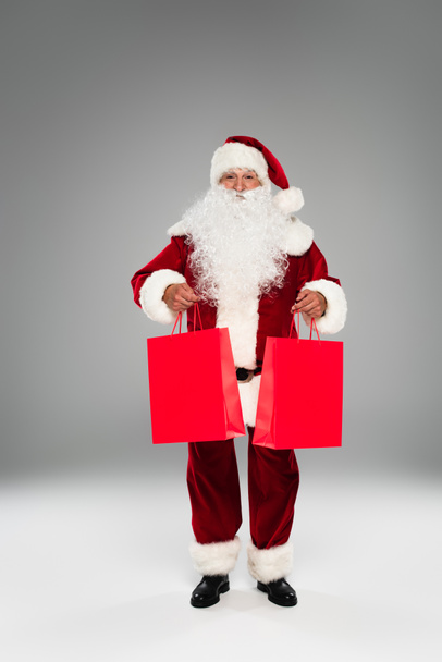 Lunghezza completa di Babbo Natale che tiene borse della spesa rosse su sfondo grigio  - Foto, immagini