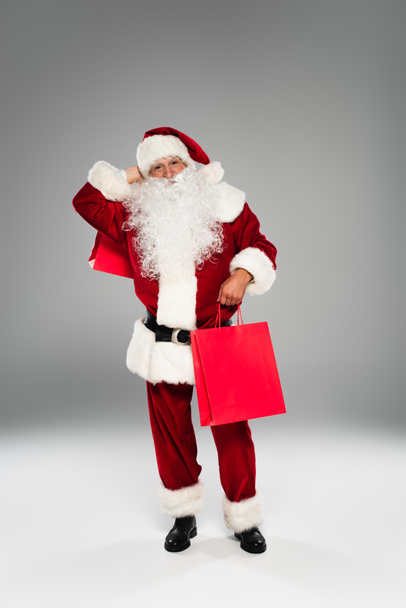 Full length of santa claus in costume holding purchases on grey background  - Φωτογραφία, εικόνα