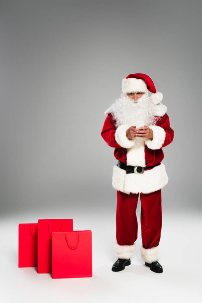 Full length of father christmas using cellphone near shopping bags on grey background  - Foto, Imagen