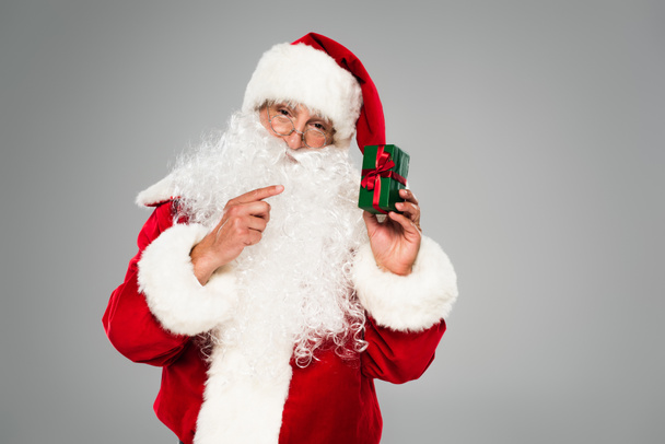 Święty Mikołaj w okularach wskazujący na małe pudełko prezentów odizolowane na szaro  - Zdjęcie, obraz
