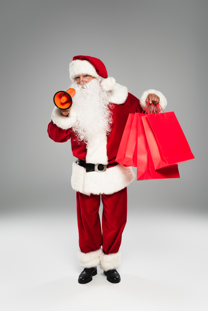 Santa claus in costume and eyeglasses holding loudspeaker and shopping bags on grey background  - Photo, Image