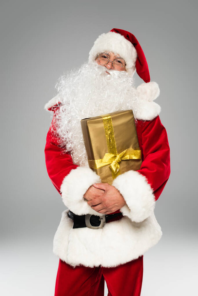 Santa claus in hat and eyeglasses holding present and looking at camera isolated on grey  - Fotografie, Obrázek