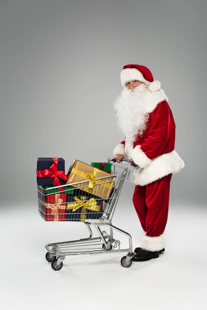Lunghezza completa di Babbo Natale in costume e occhiali in piedi vicino al carrello della spesa con regali su sfondo grigio  - Foto, immagini