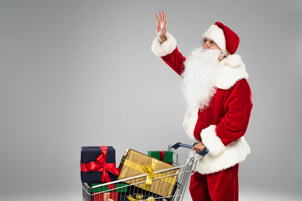 Santa claus in hat waving hand near shopping cart with presents isolated on grey  - 写真・画像