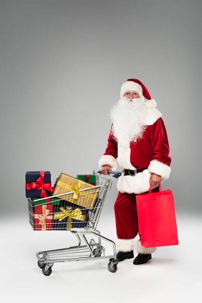 Father christmas holding shopping bags near cart with gifts on grey background  - Foto, Imagem