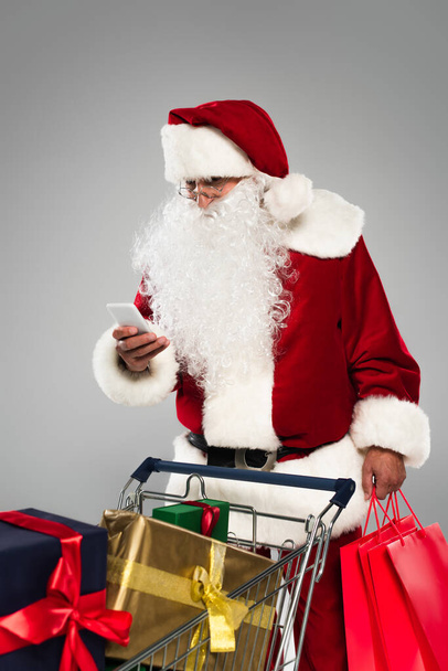 Weihnachtsmann mit Handy und Einkaufstüten in der Nähe von Geschenken im Einkaufswagen isoliert auf grau  - Foto, Bild