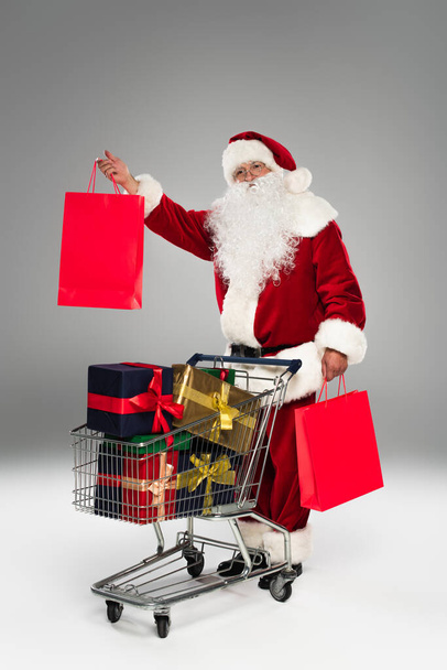 Santa claus holding shopping bags and looking at camera near presents in cart on grey background  - Photo, Image