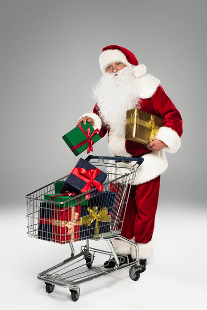Full length of santa claus in costume holding present near shopping cart on grey background  - Photo, Image