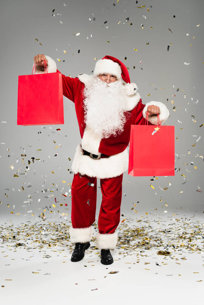 Santa claus holding red shopping bags under confetti on grey background  - Fotografie, Obrázek
