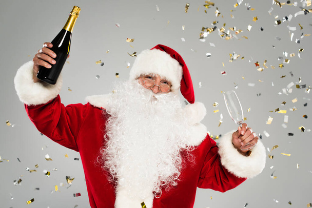 Санта-Клаус в окулярах тримає пляшку шампанського і склянку під конфетті на сірому фоні
  - Фото, зображення