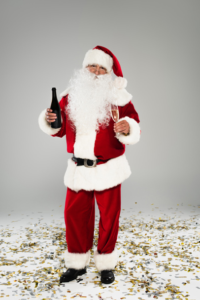 Lunghezza completa di Babbo Natale in cappello contenente bottiglia di champagne e vetro vicino coriandoli su sfondo grigio  - Foto, immagini