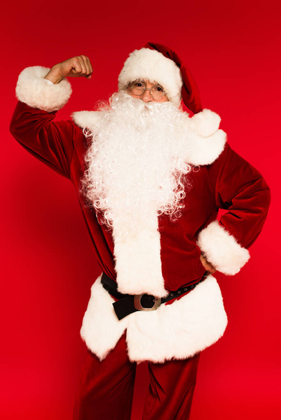 Père Noël en costume montrant le muscle sur le bras sur fond rouge  - Photo, image