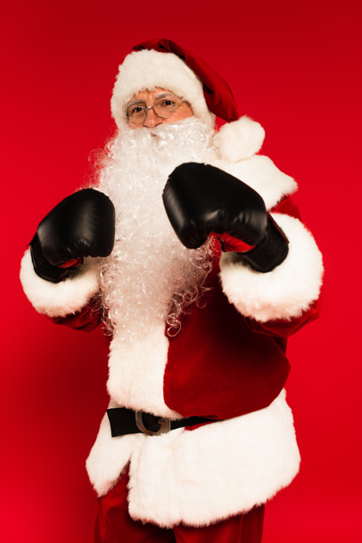 Father christmas in boxing gloves standing in fighting pose on red background  - Photo, Image
