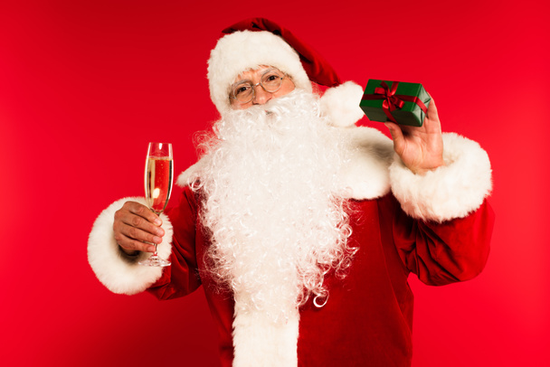 Padre Navidad celebración de copa de champán y presente sobre fondo rojo  - Foto, Imagen