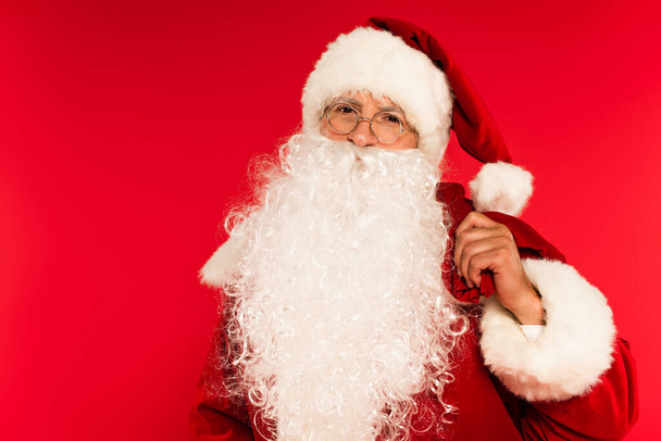 Bearded santa claus holding sack and looking at camera isolated on red  - Фото, изображение