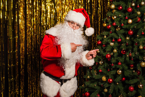 Babbo Natale punta con le dita all'albero di Natale decorato vicino ai fili di lame  - Foto, immagini