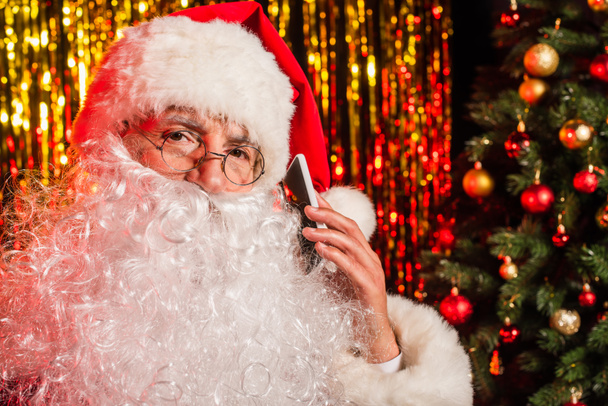 Bärtiger Weihnachtsmann in Brille spricht auf Smartphone neben verschwommenem Weihnachtsbaum und Lametta  - Foto, Bild