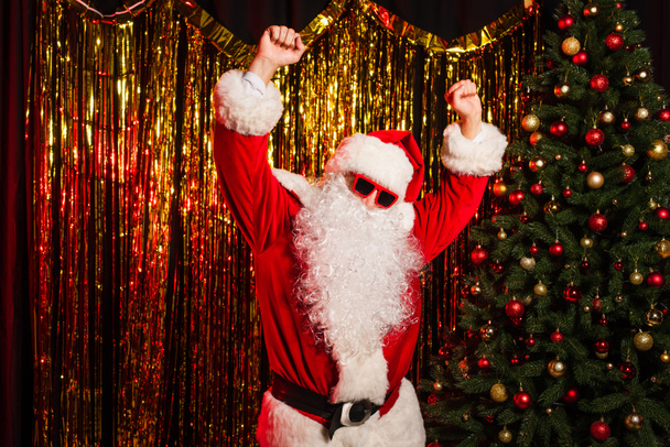 Отец Рождество в солнечных очках танцует во время вечеринки возле сосны и мишуры  - Фото, изображение