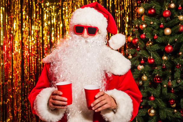 Mikulás claus napszemüveg és kalap kezében műanyag poharak közelében karácsonyfa és tinsel  - Fotó, kép