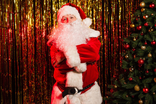 Санта-Клаус в костюме скрещивает руки и смотрит в камеру возле рождественской елки и мишуры  - Фото, изображение