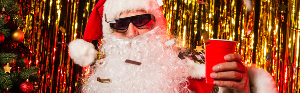 Weihnachtsmann mit Sonnenbrille hält Plastikbecher unter Konfetti in der Nähe von Lametta, Banner  - Foto, Bild