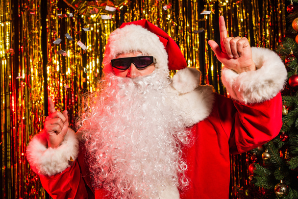 Padre Navidad en gafas de sol señalando con los dedos bajo confeti cerca de oropel festivo  - Foto, imagen