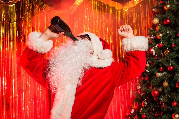 Père Noël dans les lunettes de soleil boire du champagne de bouteille près de l'arbre de Noël et tinsel  - Photo, image