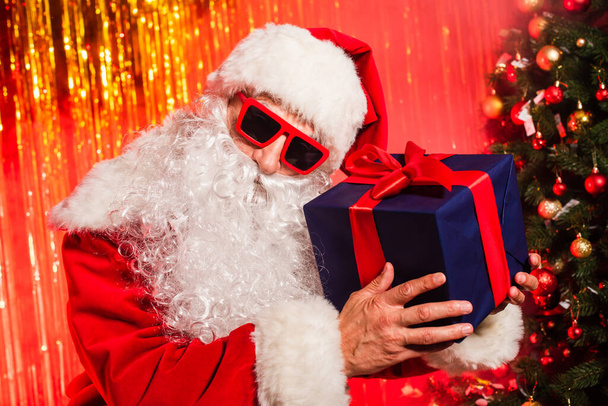 Babbo Natale in occhiali da sole e costume contenente scatola regalo durante la festa vicino all'albero di pino  - Foto, immagini