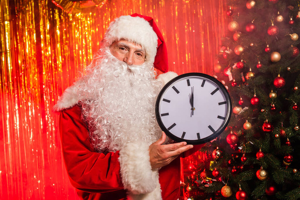 Санта-Клаус в костюме держит часы возле рождественской елки и мишуры  - Фото, изображение