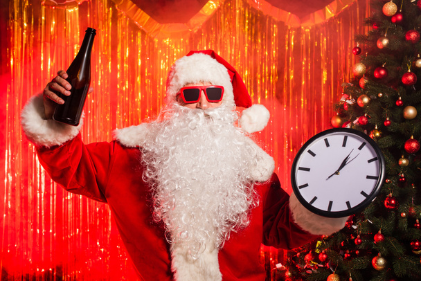 Santa Claude dans des lunettes de soleil tenant bouteille de champagne et horloge près de l'arbre de Noël  - Photo, image