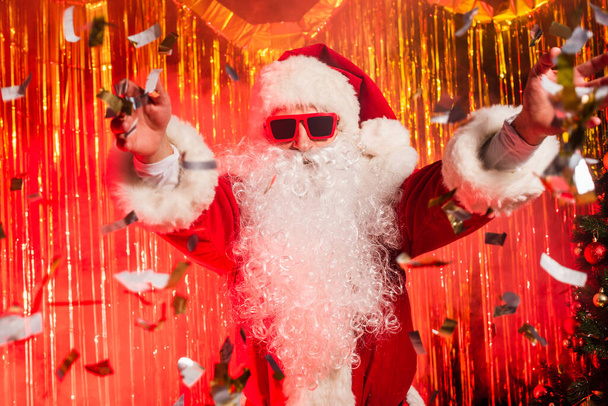 Отец Рождество в солнечных очках бросает конфетти во время вечеринки возле мишуры  - Фото, изображение