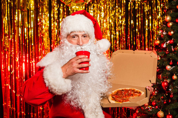 Święty Mikołaj w kostiumie trzyma plastikowy kubek i pizzę na wynos w pobliżu choinki i świecidełka  - Zdjęcie, obraz