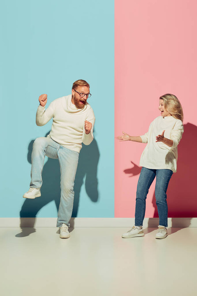 Couple of young happy man and woman dancing, having fun isolated over blue-pink studio background. Human emotions, youth, love, relationship and active lifestyle concept. Models in casual clothes - 写真・画像
