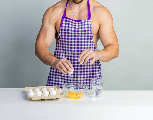 Chef bodybuilder in protective apron and cook eggs on whie kitchen table. Muscular man cooking. Healthy food and dieting. Food additives for sportsman, daily calorie diet - 写真・画像