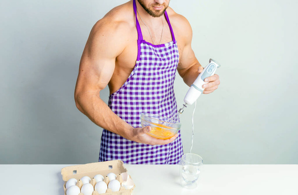Chef Bodybuilder in Schutzschürze verwendet einen Mixer, um Rührei auf einem weißen Küchentisch zu schlagen. Muskelkräftiger Mann kocht. - Foto, Bild