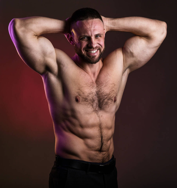 Porträt eines gut aussehenden muskulösen Bodybuilders, der vor warmem, dunklem Hintergrund posiert. Nackter muskulöser Oberkörper, Brust, Bizeps und Bauch - Foto, Bild