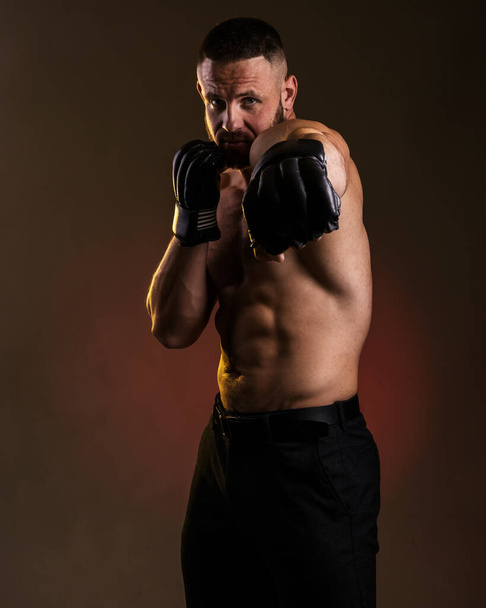 Studio portret van vechten gespierde man in zwart vechthandschoenen poseren op donkere achtergrond. Het concept van gemengde vechtsporten. Brutale bodybuilder energie en kracht boksen. - Foto, afbeelding
