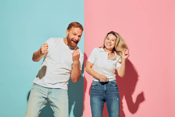 Couple of young happy man and woman dancing, having fun isolated over blue-pink studio background. Human emotions, youth, love, relationship and active lifestyle concept. Models in casual clothes - Fotografie, Obrázek