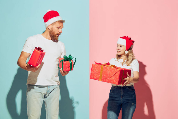 Give presents. Young charming couple wearing christmas hats and white warm sweaters holding festive boxes, smiling. Relationship, care, love and romantic, holidays, gifts, wishes, New Year concept. - Fotó, kép